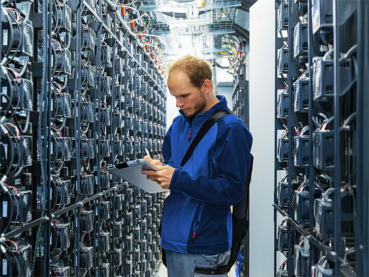 Ein Mitarbeiter steht in einer großen Batterie-Anlage und untersucht Daten auf seinem Tablet.