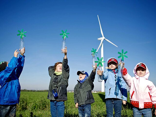Eine Gruppe Kinder stehen mit einem selbst gebastelten Windrad vor einer große Windenergie-Anlage und lachen in die Kamera.