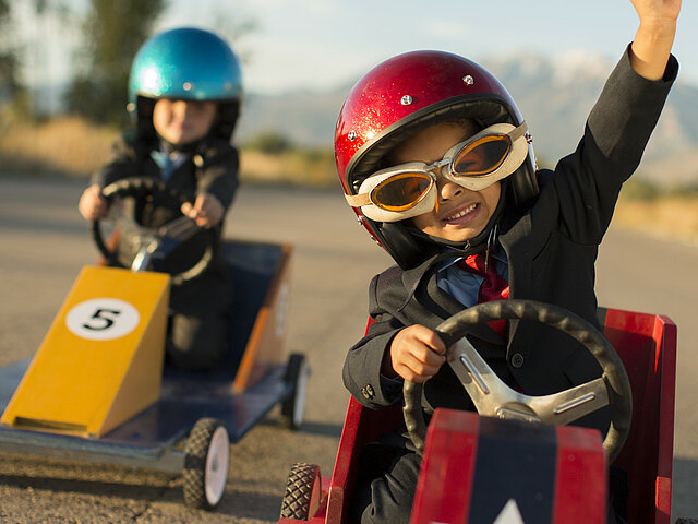 Zwei Kinder sitzen mit Rennfahrerhelm in einer Seifenkiste und fahren eine Straße entlang.