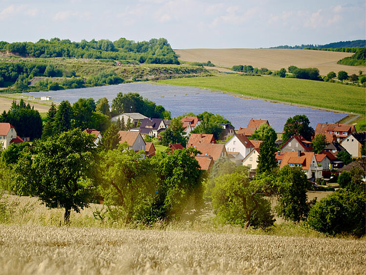Hinter einem Feld mit reifem Getreide ist eine kleine Gmeinde zu sehen, dahinter steht eine PV-Anlage.