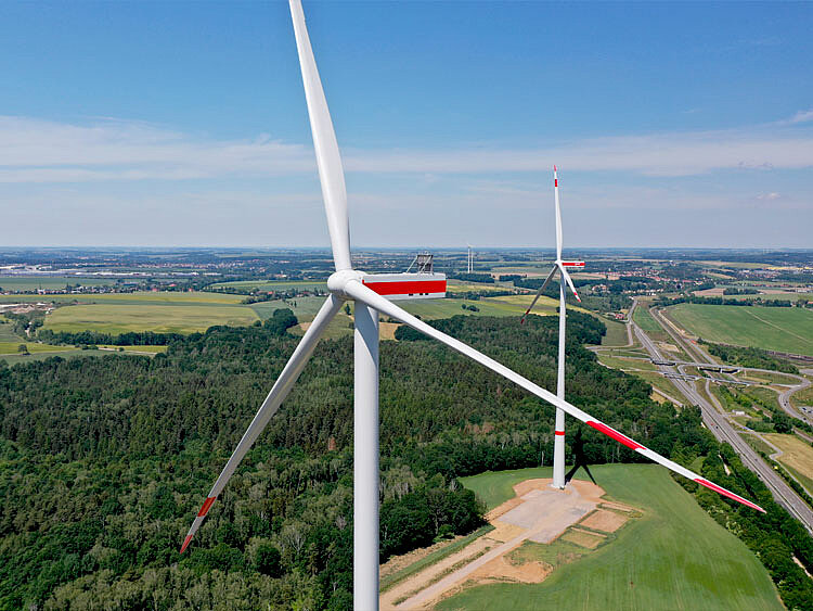 Zwei Windenergie-Anlage stehen im Vordergrund. Dahinter erstreckt sich eine weite Landschaft.