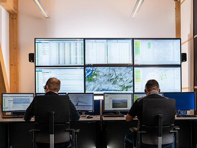 Zwei Mitarbeiter einer Leitwarte sitzen vor einer Wand an Bildschirmen.