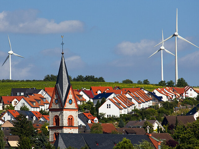 Kirchturm mit Dorfbebauung im Vordergrund mit drei Windenergieanlagen im Weinberg im Hintergrund.