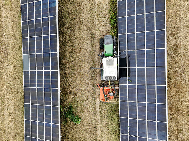 Blick von Oben: Ein Traktor mäht die Grasfläche zwischen zwei Solartischen in einem Freichflächensolarpark.