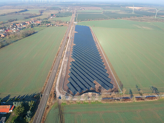 Der Solarpark Zernitz wird von der winterlichen Sonne beschnienen. Der Park liegt an einer Bahnstrecke, die Bäume am Rand haben kein Laub. Auf den Feldern ringsum ist spräliches Grün zu erkennen.