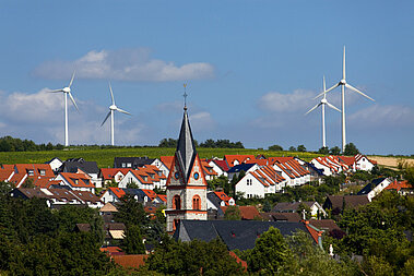 Kirchturm mit Dorfbebauung im Vordergrund mit vier Windenergieanlagen im Weinberg im Hintergrund.