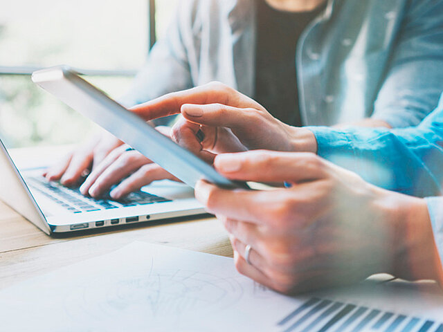 Zwei Personen, von denen nur die Arme und Oberkörper zu sehen sind, sitzen gemeinsam vor einem Laptop.
